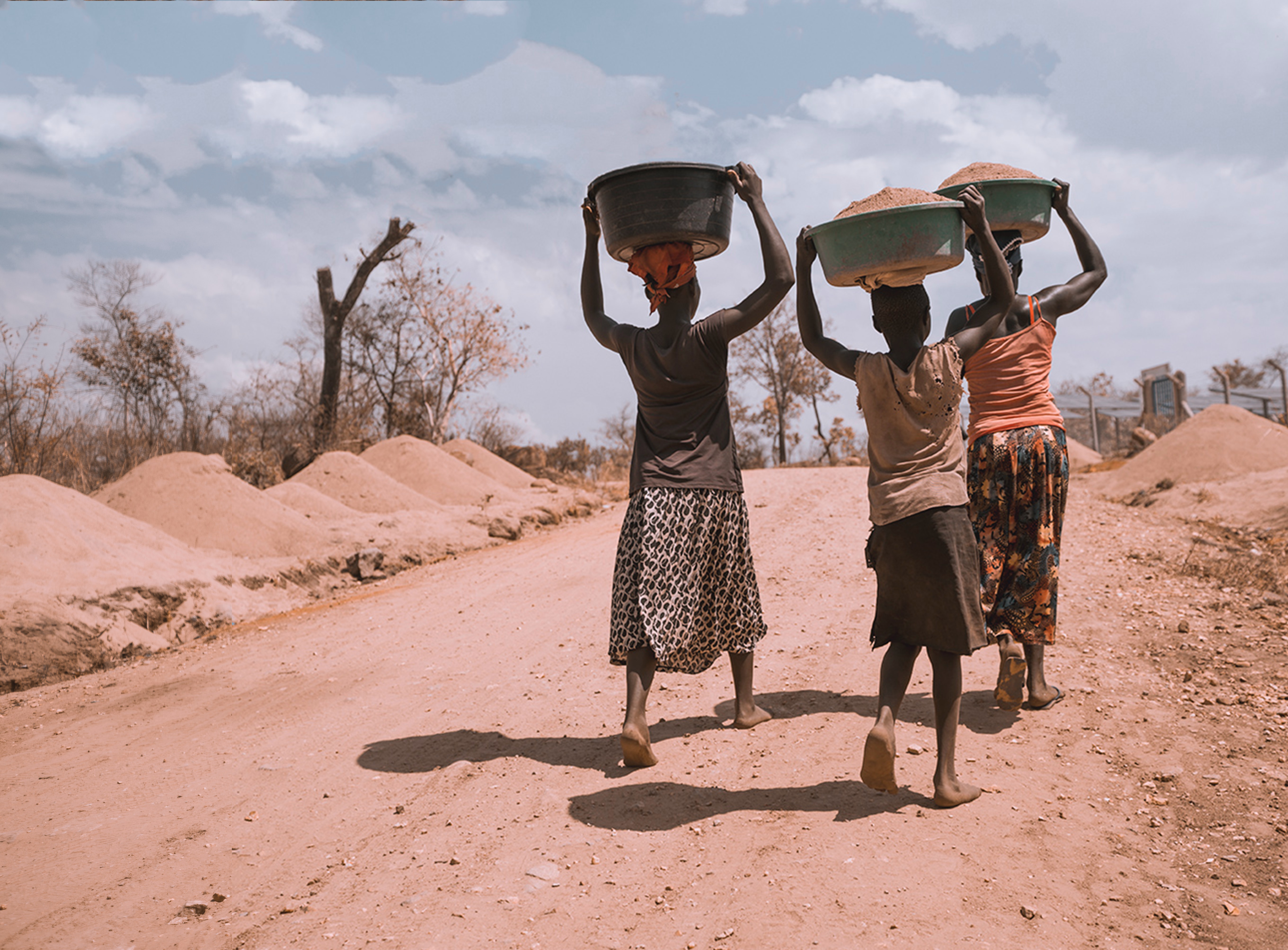 Three people walking down a dirt road with large buckets on their heads.