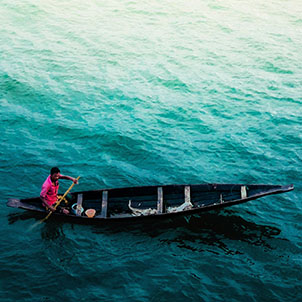 Stock image of a Bangladeshi man in a red shirt paddling a traditional canoe on blue water